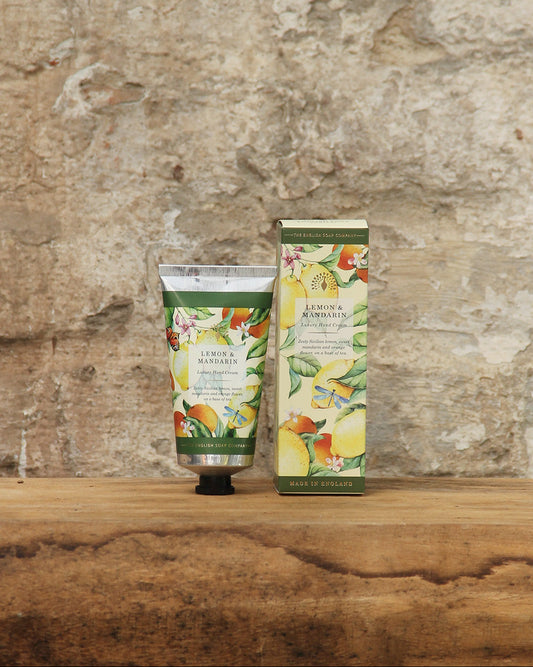 Eine Tube und eine Schachtel der ESC | 'Lemon Mandarin' Handcreme 75 ml von The English Soap Company mit lebhaften botanischen Motiven ruhen auf Holz vor einer Steinwand - eine stilvolle Kulisse für diese luxuriöse vegane Handpflege.
