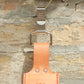 Die Niwaki | 'Belt Clip' Praktische Halterung für Niwaki Single Holster, eine tan Lederschlaufe mit japanischer Prägung und silbernem Haken, hängt als Gürtelbefestigung an einer hellen Ziegelwand.