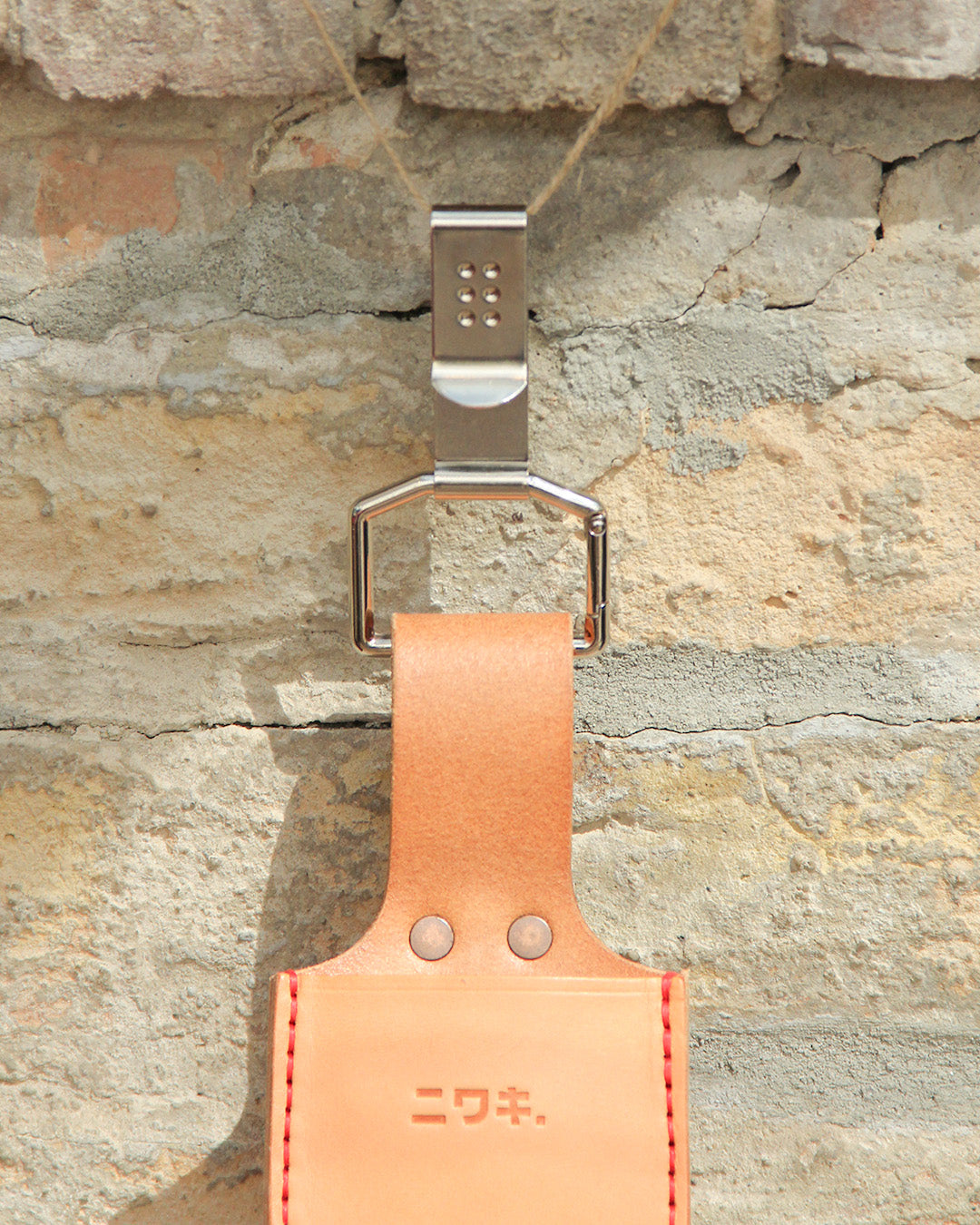 Die Niwaki | 'Belt Clip' Praktische Halterung für Niwaki Single Holster, eine tan Lederschlaufe mit japanischer Prägung und silbernem Haken, hängt als Gürtelbefestigung an einer hellen Ziegelwand.