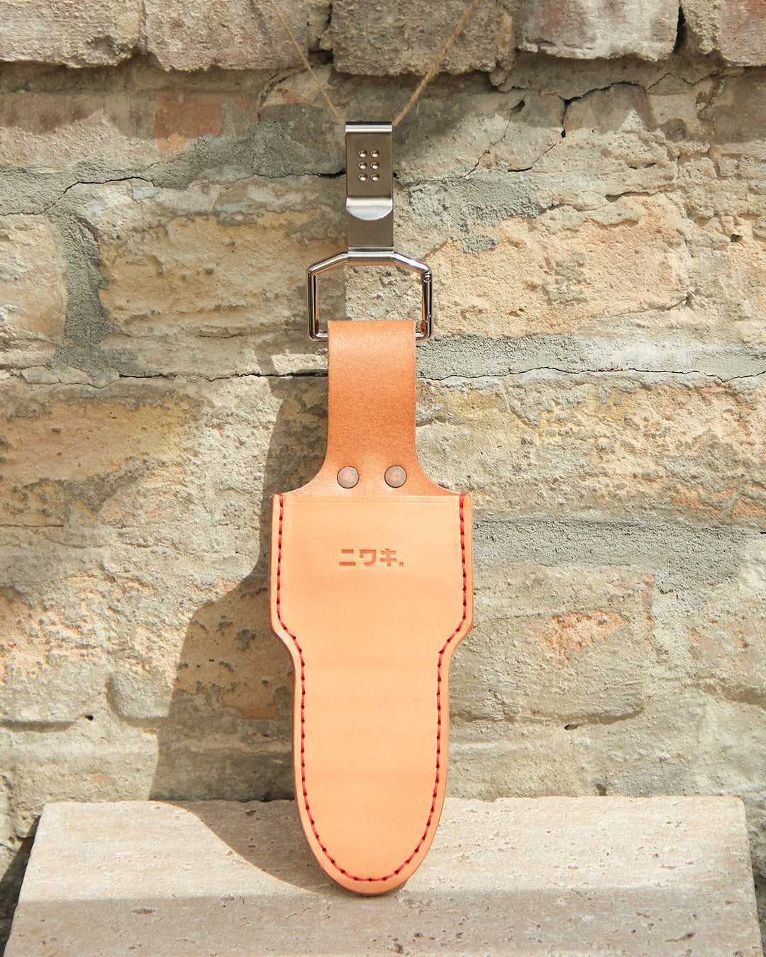 Eine Niwaki | 'Belt Clip' Praktische Halterung für Niwaki Single Holster aus hellbraunem Leder mit roten Nähten und japanischer Textprägung auf der Vorderseite hängt an einem Metallhaken vor einer strukturierten Steinwand.
