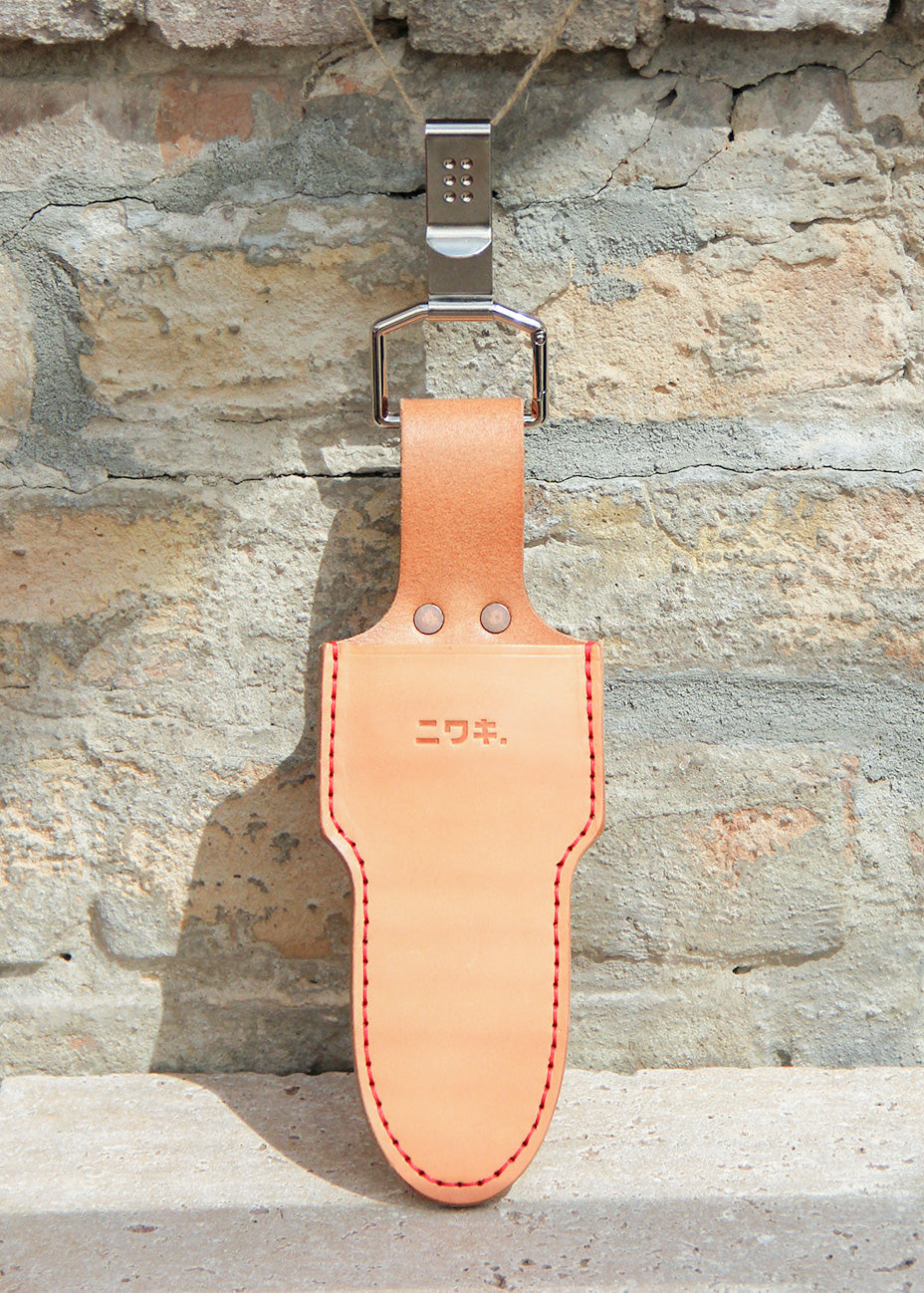 Ein hellbraunes Lederholster, das „Single Holster“ Lederschutzhülle für Schere von Niwaki, mit auffälligen roten Nähten, hängt an einem Metallhaken vor einer strukturierten Backsteinwand. Auf der Vorderseite dieser Scheren-Schutzhülle sind japanische Schriftzeichen eingeprägt, die dem markanten Design eine elegante Note verleihen.