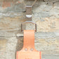 Ein „Belt Clip“ von Niwaki – ein praktischer Halter aus Leder mit japanischer Aufschrift – hängt an einem Metallhaken, der an einer strukturierten, hellen Ziegelwand befestigt ist.