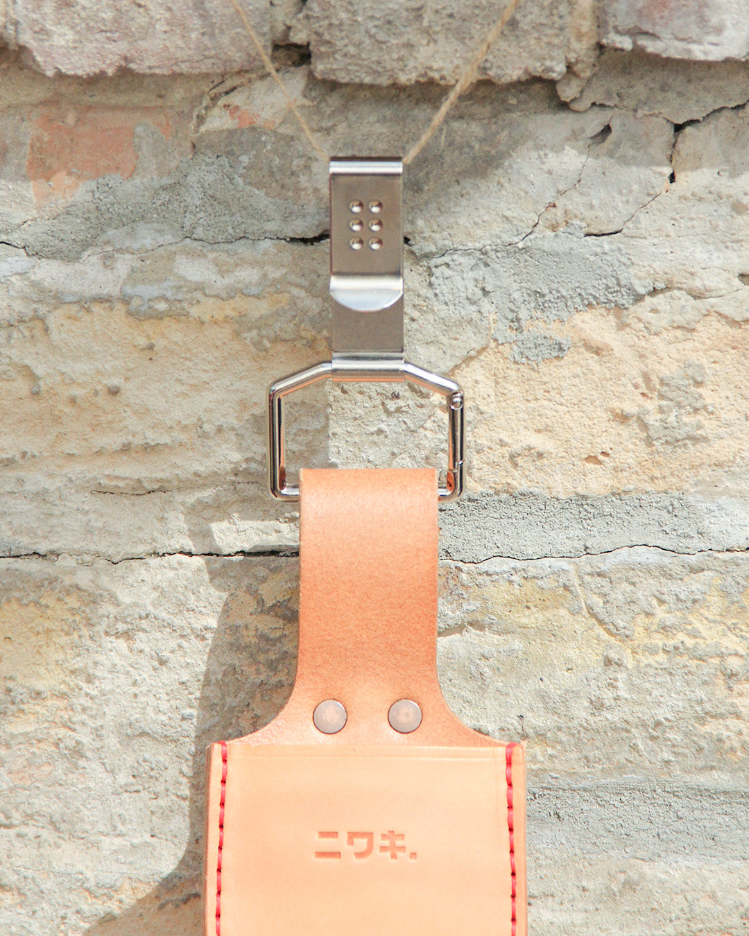Ein „Belt Clip“ von Niwaki – ein praktischer Halter aus Leder mit japanischer Aufschrift – hängt an einem Metallhaken, der an einer strukturierten, hellen Ziegelwand befestigt ist.