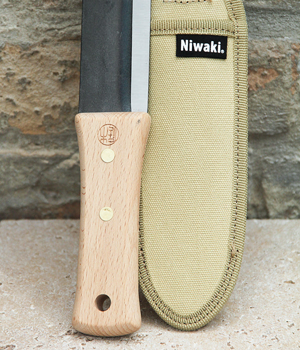 Ein Niwaki | Das multifunktionale Pflanzmesser „Hori Hori“ mit Holzgriff ist neben einer beigen Schutzhülle mit Niwaki-Logo auf einer Steinoberfläche vor einer rauen Steinwand zu sehen – ideal für präzise Gartenarbeiten.