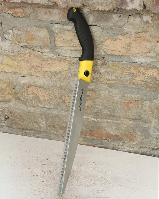 Eine große Niwaki GR 270 Baumsäge mit schwarz-gelbem Griff steht aufrecht, mit dem Sägeblatt nach unten, vor einer rauen Ziegelwand auf einer weißen Fläche mit Holzspänen. Die Klinge trägt das Niwaki-Logo.
