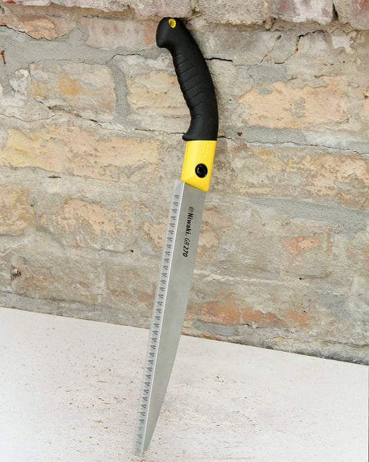 Eine Niwaki „GR 270 Pruning Saw“ Astsäge mit Schutzhülle, deren schwarz-gelber Griff an eine japanische Baumsäge erinnert, lehnt an einer rauen Ziegelwand. Ihr Sägeblatt verfügt über scharfe Zähne für präzise Schnittarbeiten, und auf der weißen Fläche darunter liegen Holzspäne verstreut.