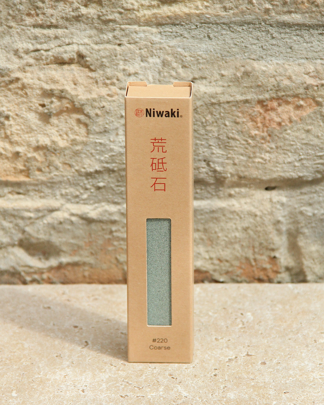 Der Niwaki 'Sharping Stone' Schleifstein grob (#220 Coarse) in einer Pappschachtel mit englischem und rotem japanischem Text steht aufrecht auf dem Stein - ideal zum Schärfen von Gartengeräten und für verschiedene Schleifstufen.