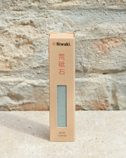 Ein rechteckiger Niwaki | „Sharping Stone“ Schleifstein grob von Niwaki, verpackt in einer hellbraunen Schachtel mit der Aufschrift „Coarse #220“, steht aufrecht auf einer Steinoberfläche vor einer strukturierten Steinwand – ideal zum Schärfen von Gartengeräten in verschiedenen Schleifstufen.
