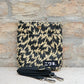 Eine kompakte, beige-schwarze Tasche mit Zickzackmuster von Niwaki im Stil „Eley Kishimoto X Niwaki Flash“ mit Druckknopfverschluss lehnt an einer Backsteinmauer. Davor liegt ein schwarzer Riemen mit Metallklammern. Auf dem Boden der Tasche ist auf einem schwarzen Aufnäher weißer japanischer Text zu sehen.