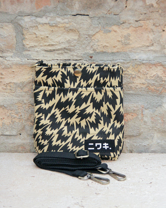 Eine kompakte, beige-schwarze Tasche mit Zickzackmuster von Niwaki im Stil „Eley Kishimoto X Niwaki Flash“ mit Druckknopfverschluss lehnt an einer Backsteinmauer. Davor liegt ein schwarzer Riemen mit Metallklammern. Auf dem Boden der Tasche ist auf einem schwarzen Aufnäher weißer japanischer Text zu sehen.