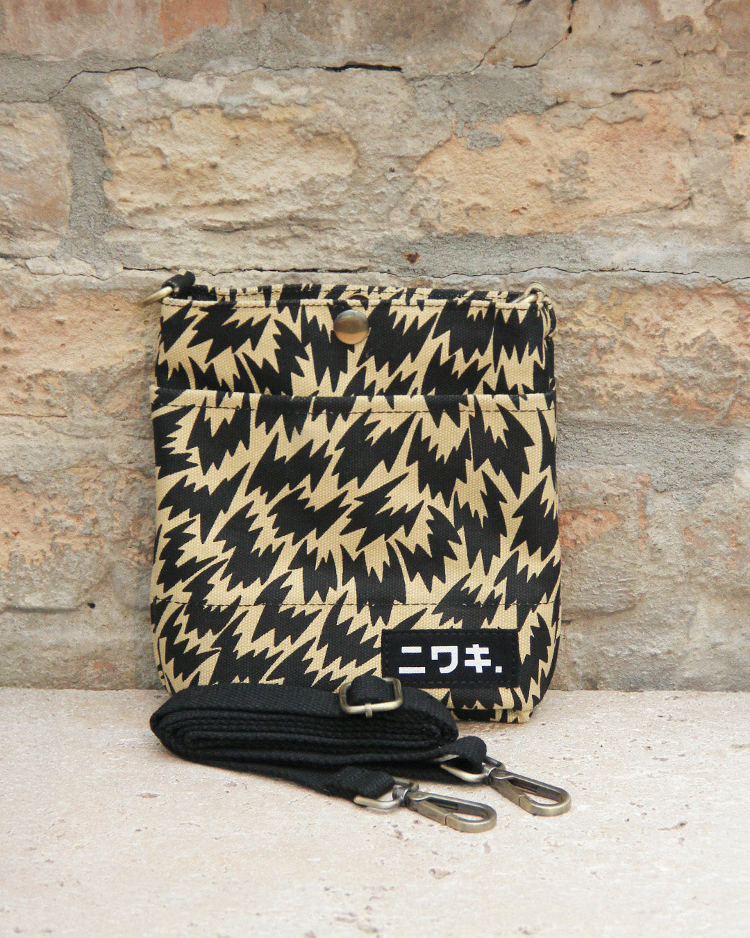 Eine kleine Niwaki Kantan-Umhängetasche im „Eley Kishimoto X Niwaki Flash“-Stil lehnt an einer Backsteinmauer und präsentiert ihr auffälliges Zickzack-Muster in Schwarz und Hellbraun. Diese wasserabweisende Canvas-Tasche verfügt über einen schwarzen Riemen mit Metallclips an der Vorderseite der Tasche. Weiße japanische Schriftzeichen auf einem schwarzen Aufnäher verleihen dem Modell einen zusätzlichen Hauch von Stil der Marke Niwaki.