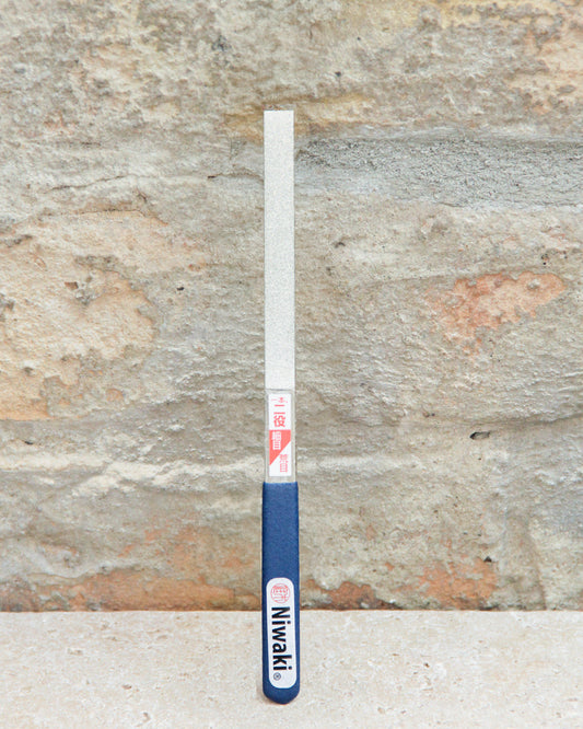 Eine doppelseitige Diamantfeile mit blauem Griff von Niwaki, beschriftet mit „Niwaki“, steht aufrecht vor einer strukturierten Steinwand – ideal zum Schärfen von Gartenscheren mit der hochwertigen doppelseitigen Diamantfeile „Twin Diamond File“ von Niwaki.