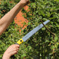 Jemand benutzt die Niwaki | 'GR 270 Pruning Saw' Astsäge mit Schutzhülle von Niwaki mit ihrem gelb-schwarzen Griff, um bei sonnigen Gartenarbeiten im Freien einen Ast zu schneiden.