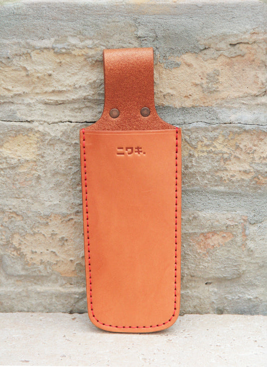 Eine Niwaki | „Saw Holster“ – eine hochwertige Schutzhülle aus hellbraunem Leder für eine Klappsäge, mit roten Nähten und geprägten japanischen Schriftzeichen auf der Vorderseite, von Niwaki gefertigt, um Gartengeräte optimal zu schützen, hängt elegant an einer hellen Ziegelwand.