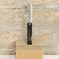 Ein Niwaki „SK Higonokami Folding Knife“ Klappmesser (171 mm) mit japanischen Schriftzeichen auf dem Griff steht aufrecht auf einem Stück Wellpappe vor dem Hintergrund einer strukturierten Steinwand. Dieses traditionelle Niwaki-Taschenmesser verkörpert klassische japanische Handwerkskunst und Stil.