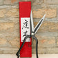 Ein Paar Niwaki "Sentei Topiary Clippers" mit schwarzen Griffen und scharfen silbernen Klingen ruht vor einer strukturierten Ziegelwand, vor einer hohen roten Schachtel mit vertikaler japanischer Kalligrafie auf einem weißen Etikett.