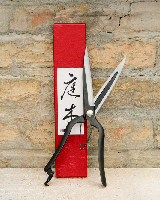 Ein Paar Niwaki "Sentei Topiary Clippers" mit schwarzen Griffen und scharfen silbernen Klingen ruht vor einer strukturierten Ziegelwand, vor einer hohen roten Schachtel mit vertikaler japanischer Kalligrafie auf einem weißen Etikett.