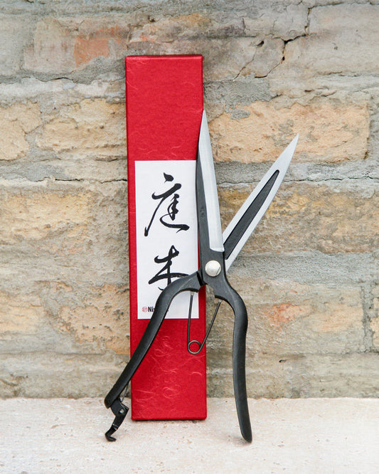 Niwaki | „Sentei Topiary Clippers“ – Buchsbaumschere in Schwarz, aufrecht vor einer strukturierten Backsteinmauer präsentiert, zusammen mit der markanten roten Schachtel mit japanischer Kalligraphie.