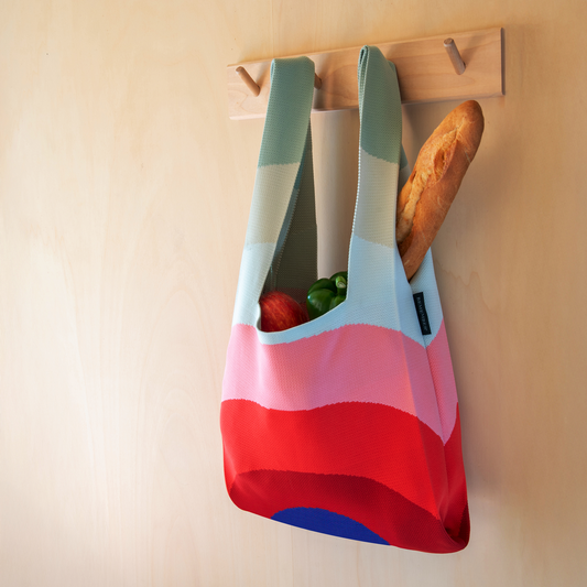 Die Remember Stricktasche 'Indigo' aus recyceltem Polyester hängt an einem hölzernen Wandhaken mit Lebensmitteln darin - ein stilvolles Accessoire von Remember für die täglichen Besorgungen.