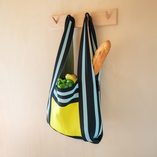 Die Remember Stricktasche 'Luce' aus recyceltem Polyester zeigt ein modernes Streifendesign in Schwarz, Blau und Gelb, hängt am Holzhaken und bietet Platz für Baguette sowie frisches Gemüse.