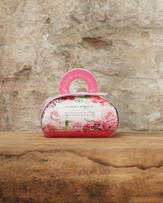 Auf einer rustikalen Holzfläche vor einer Steinmauer sitzt The English Soap Company ESC | 'Summer Rose' blumig duftende pflanzliche Handseife in einer pinken, plastikfreien Geschenkbox mit Rosendesign, Made in England.