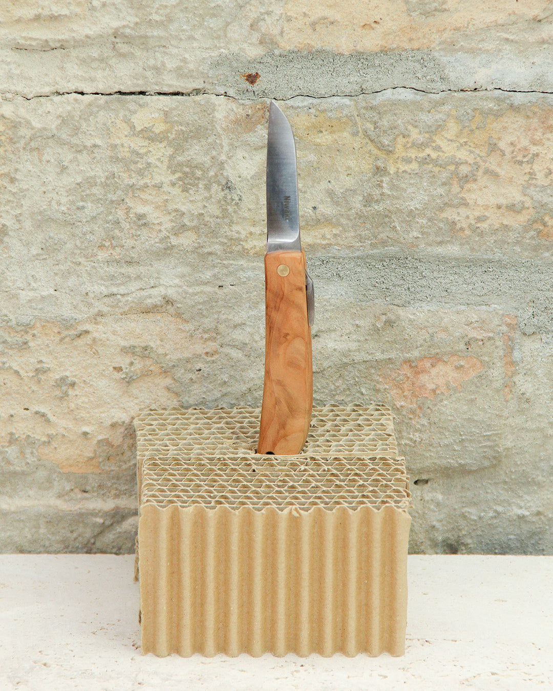 Ein Niwaki | „Kotoh X Niwaki Knife“ Taschenmesser mit Lederschutzhülle steht aufrecht, mit geöffneter Klinge in einen Block aus Wellpappe gesteckt. Der Hintergrund zeigt eine strukturierte Steinwand und vermittelt so die Präzision und Eleganz, die oft mit dem Bonsai-Schnitt und der fachkundigen Gartenarbeit von Niwaki in Verbindung gebracht werden.