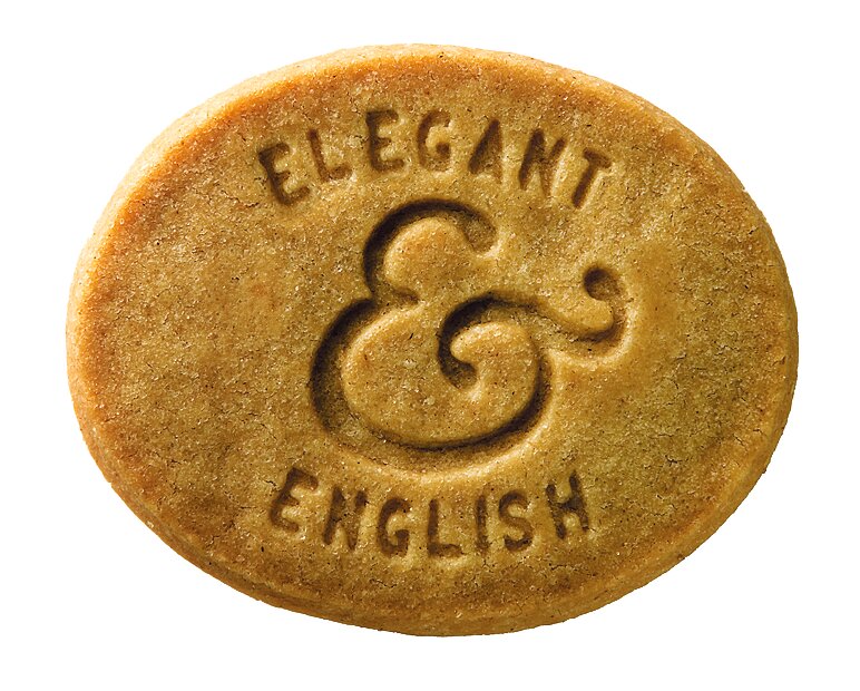 Ein runder, goldbrauner Butterkeks von Lady Joseph mit der Prägung ELEGANT & ENGLISH und einem dekorativen kaufmännischen Und-Zeichen ziert die 130 g-Packung Elegant & English | Butterkekse mit Zitrone & Ingwer auf weißem Hintergrund.