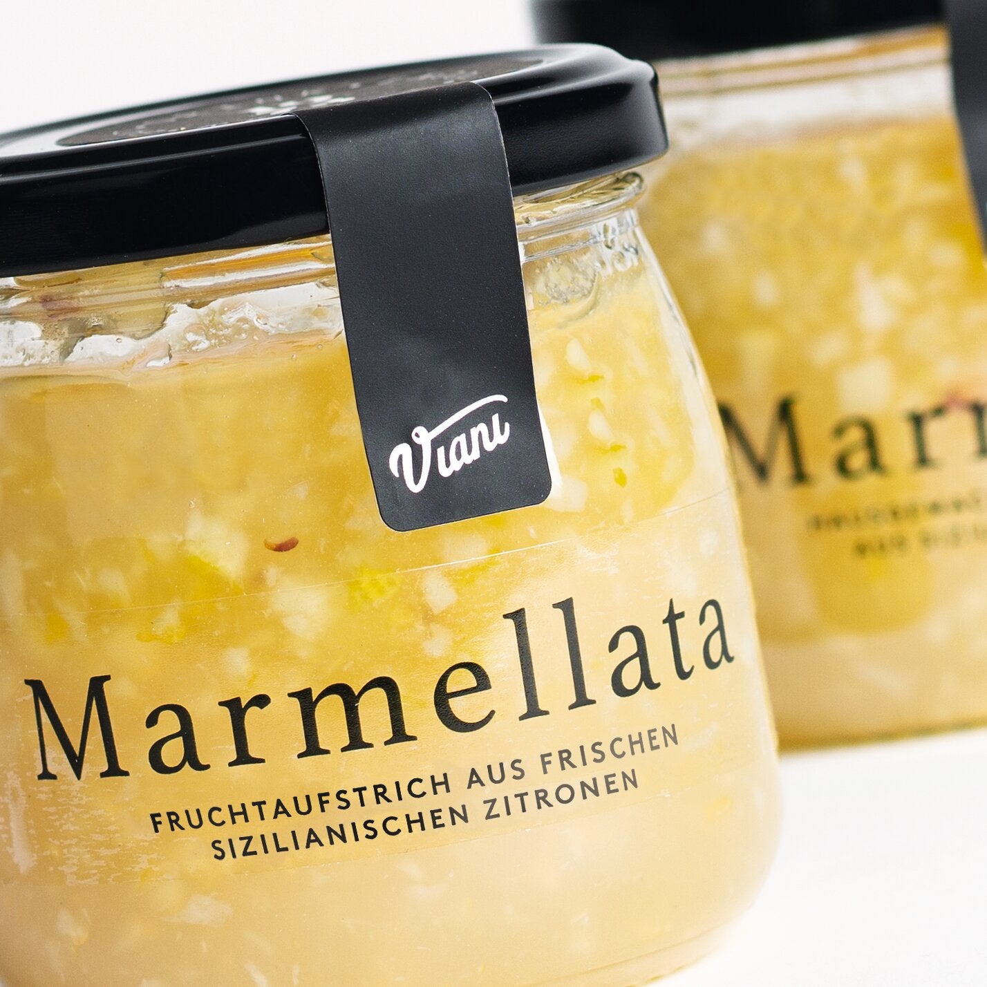 Nahaufnahme von zwei Viani-Gläsern mit der Aufschrift "Marmellata di Limoni" Fruchtaufstrich aus sizilianischen Zitronen 210 g, mit deutschem Text, der diesen sizilianischen Zitronenfruchtaufstrich beschreibt.