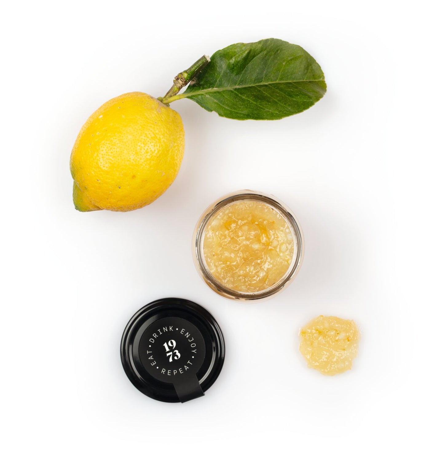 Eine ganze Zitrone mit einem Blatt, ein geöffnetes Glas Viani | 'Marmellata di Limoni' Fruchtaufstrich aus sizilianischen Zitronen 210 g mit schwarzem Deckel und ein Klecks Fruchtaufstrich auf weißem Grund.