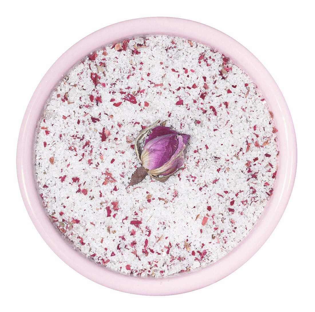 In einer rosafarbenen Schale befindet sich das Meersalz Fleur de Sel La Vie en Rose" von Sal de Ibiza, das mit Rosenblättern vermengt und in der Mitte mit einer einzelnen getrockneten Rosenknospe gekrönt ist.