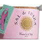 Ein rosafarbenes Keramikgefäß mit Korkdeckel, weißem Löffel und goldenem Sonnenlogo enthält Sal de Ibiza 'Fleur de Sel La Vie en Rose' Meersalz mit Rosenblättern (140 g). Ein grünes Etikett mit rosa Schleife macht auf das Thema Brustkrebs aufmerksam.