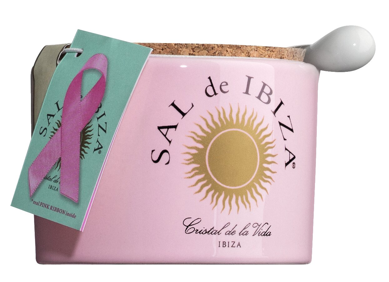 Ein rosafarbenes Keramikgefäß mit Korkdeckel, weißem Löffel und goldenem Sonnenlogo enthält Sal de Ibiza 'Fleur de Sel La Vie en Rose' Meersalz mit Rosenblättern (140 g). Ein grünes Etikett mit rosa Schleife macht auf das Thema Brustkrebs aufmerksam.