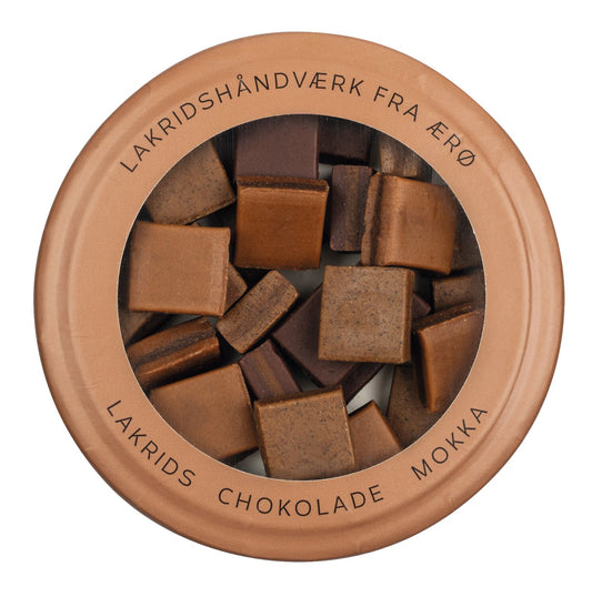 In einer runden Dose mit durchsichtigem Deckel befinden sich quadratische Stücke brauner Lakritzschokolade. Am Rand des Deckels ist in schwarzen Buchstaben die Aufschrift „LAKRIDSHÅNDVÆRK FRA ÆRØ LAKRIDS CHOKOLADE MOKKA“ zu lesen, die die handwerkliche Qualität von Hattesens Lakritzkonfekt widerspiegelt. Diese 125-g-Köstlichkeit, Hattesens Lakritzkonfekt | Lakritz, Schokolade & Mokka, vereint auf meisterhafte Weise traditionelle dänische Handwerkskunst mit unwiderstehlichem Lakritzkonfekt in jedem Bissen.