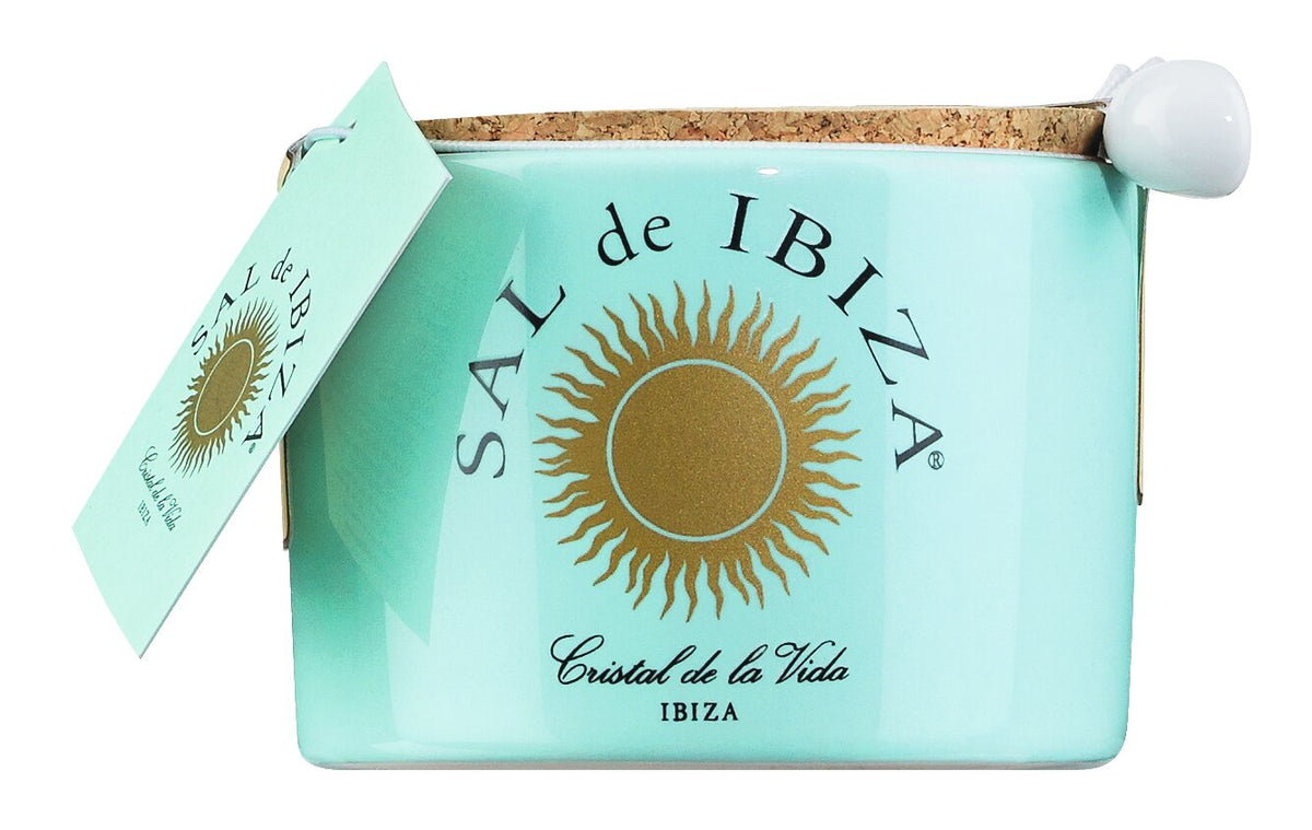 Ein türkisfarbenes Keramikgefäß mit „Fleur de Sel“-Meersalz von Sal de Ibiza, 150 g, verziert mit einem goldenen Sonnenlogo, einem Korkdeckel, einem beigefügten Löffel und einem passenden Etikett. Unterhalb der Sonne erscheint der Schriftzug „Cristal de la Vida Ibiza“, der das hochwertige Fleur de Sel-Meersalz hervorhebt.