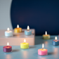 Mehrere kleine, farbenfrohe Remember Teelicht Set 'Sydney' Kerzen aus umweltfreundlichem, veganem Stearinwachs werden auf einer blauen Fläche mit verschwommenem navy-beigem Hintergrund angezündet und schaffen eine gemütliche und heitere Atmosphäre.