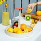 Ein rundes Korallentablett 'Nana' von Remember enthält Zitronen, Orangen und bunte Gläser. Eine Hand presst eine Zitrone über einem Getränk aus, daneben stehen ein Krug mit Zitronengetränk und ein Glas Limonade.