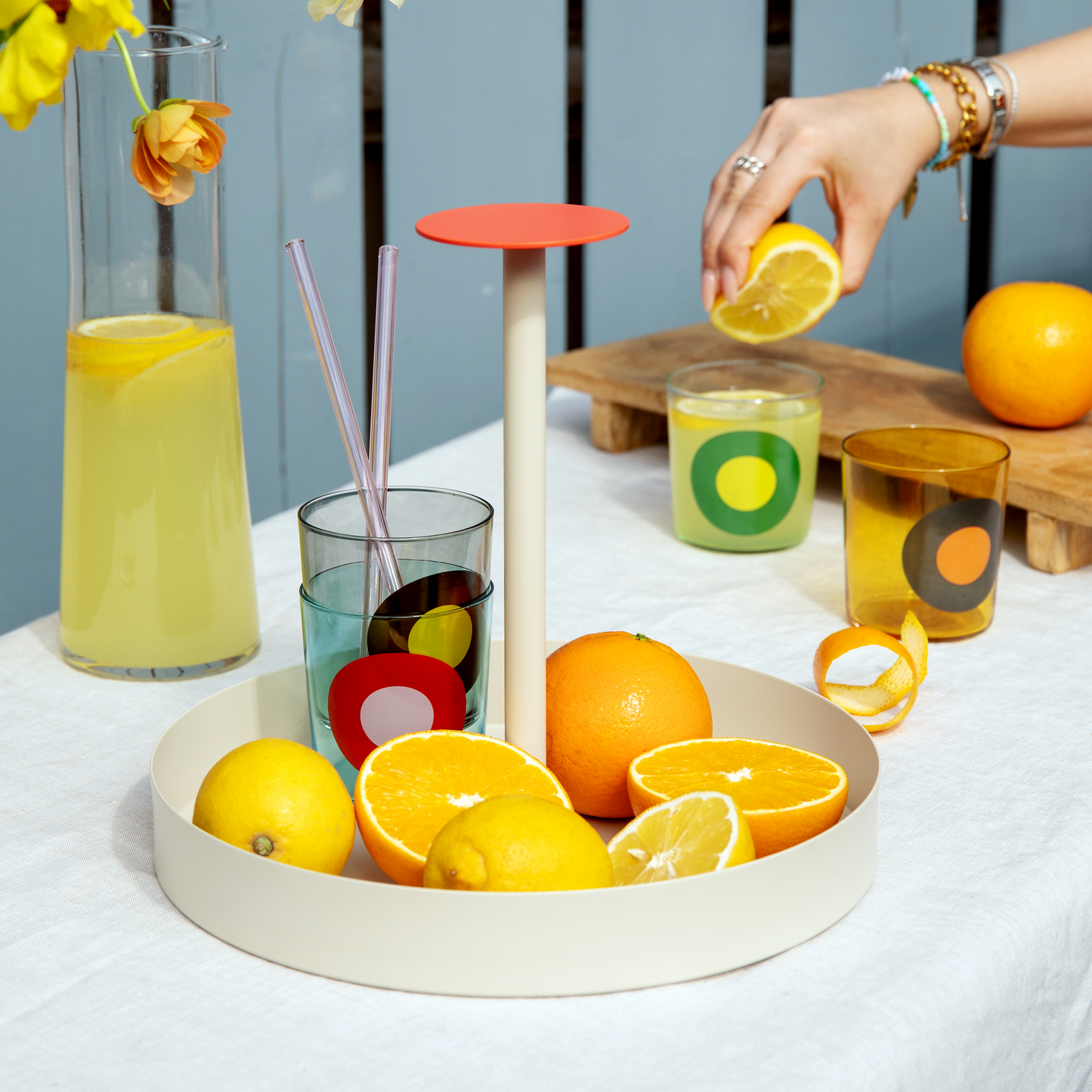 Ein rundes Korallentablett 'Nana' von Remember enthält Zitronen, Orangen und bunte Gläser. Eine Hand presst eine Zitrone über einem Getränk aus, daneben stehen ein Krug mit Zitronengetränk und ein Glas Limonade.