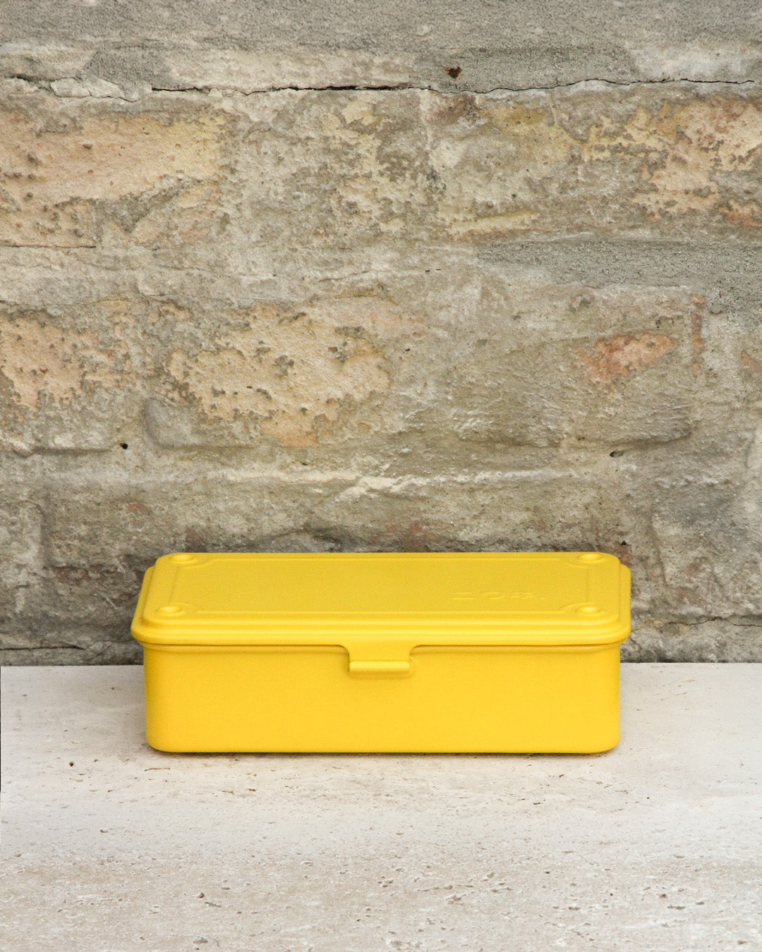 Eine geschlossene, rechteckige gelbe Niwaki 'T-Type Toolbox' Werkzeugbox (19 x 9 x 5 cm) ruht auf einer hellen Fläche mit einer strukturierten Steinwand im Hintergrund.