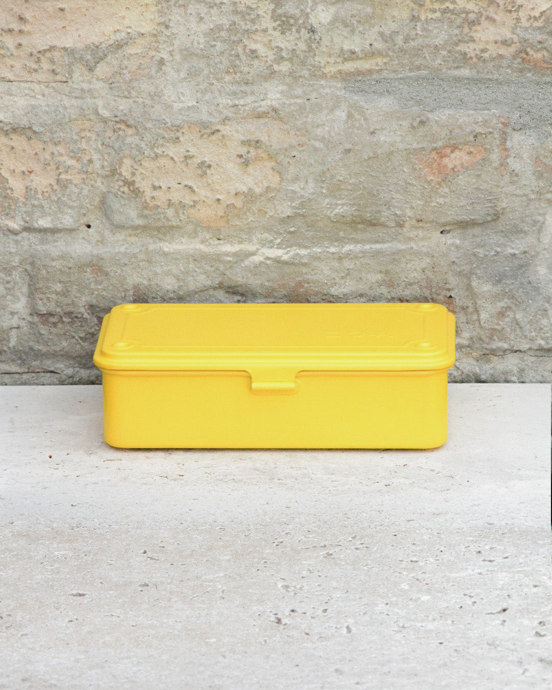 Eine gelbe, rechteckige Niwaki-Werkzeugbox „T-Type Toolbox“ aus Kunststoff (19 x 9 x 5 cm) mit Deckel steht auf einer hellen Betonfläche vor dem Hintergrund einer strukturierten Steinwand.