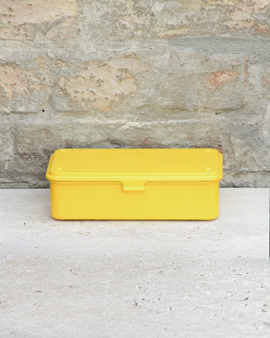 Eine gelbe, rechteckige Niwaki-Werkzeugbox „T-Type Toolbox“ aus Kunststoff (19 x 9 x 5 cm) mit Deckel steht auf einer hellen Betonfläche vor dem Hintergrund einer strukturierten Steinwand.