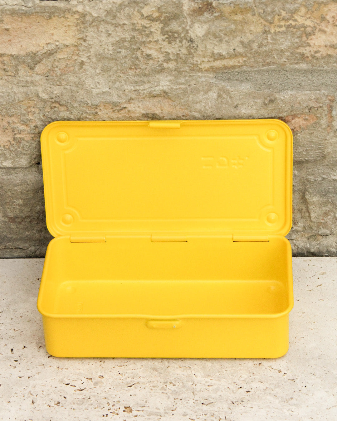 Eine gelbe Niwaki 'T-Type Toolbox' Werkzeugbox (19 x 9 x 5 cm), offen und leer, ruht auf einer hellen Steinoberfläche vor einer strukturierten Steinwand.