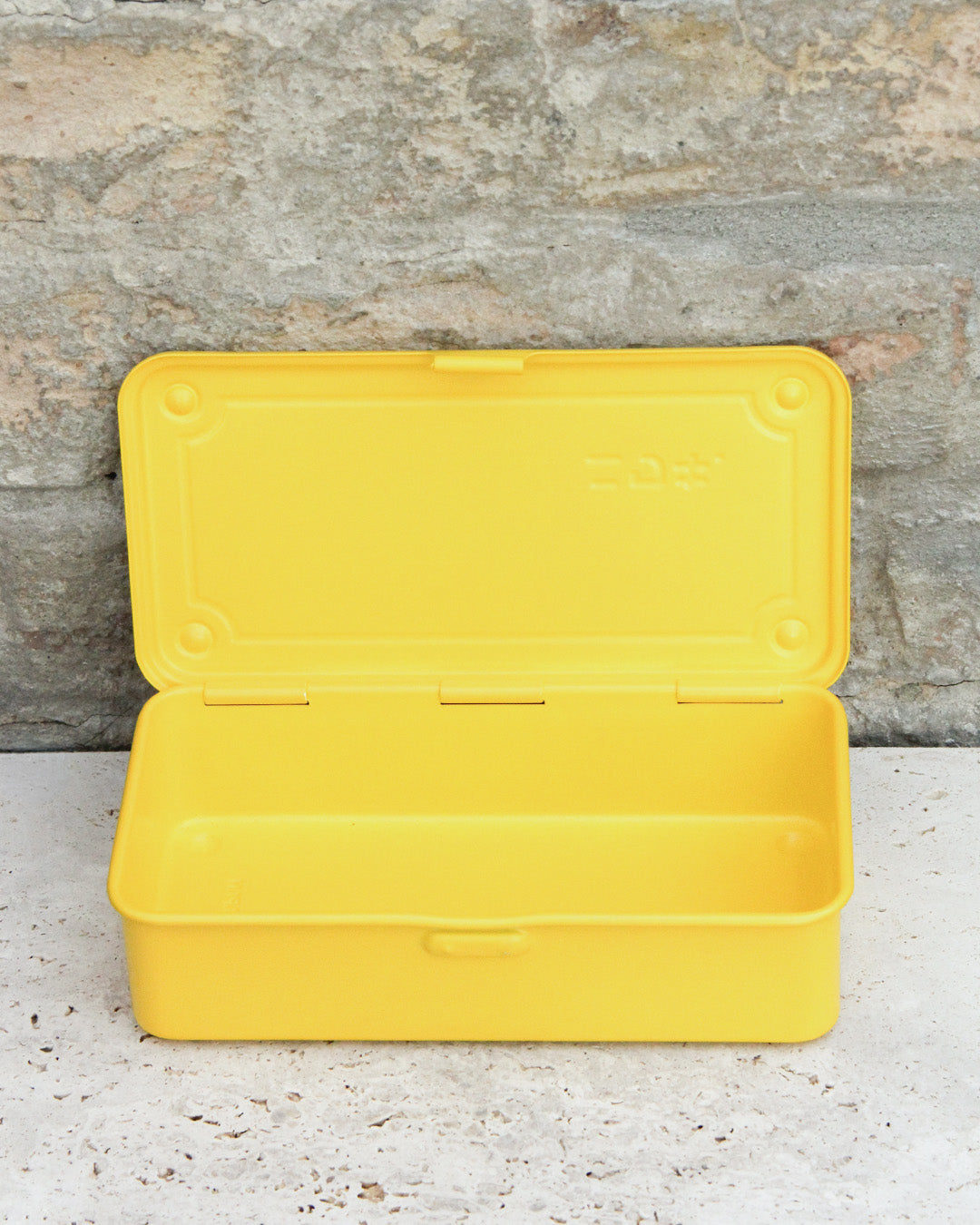 Eine gelbe Niwaki „T-Type Toolbox“-Werkzeugbox (19 x 9 x 5 cm) aus Kunststoff steht mit geöffnetem Deckel auf einer hellen Steinfläche vor einer Steinmauer.
