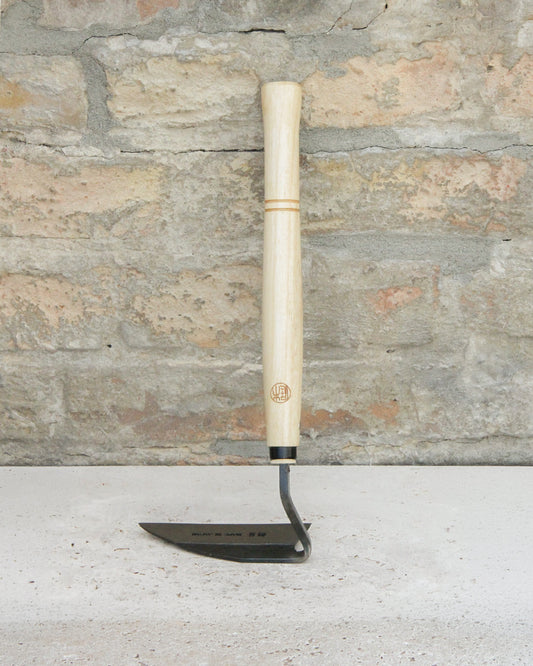 Die Niwaki | „Weeding Hoe“ Jäthacke für Rechts- und Linkshänder mit einem leichten Holzstiel und einer flachen, abgewinkelten Metallklinge lehnt aufrecht an einer strukturierten Steinmauer – ideal, um mühelos Unkraut in Ihrem Garten zu entfernen.