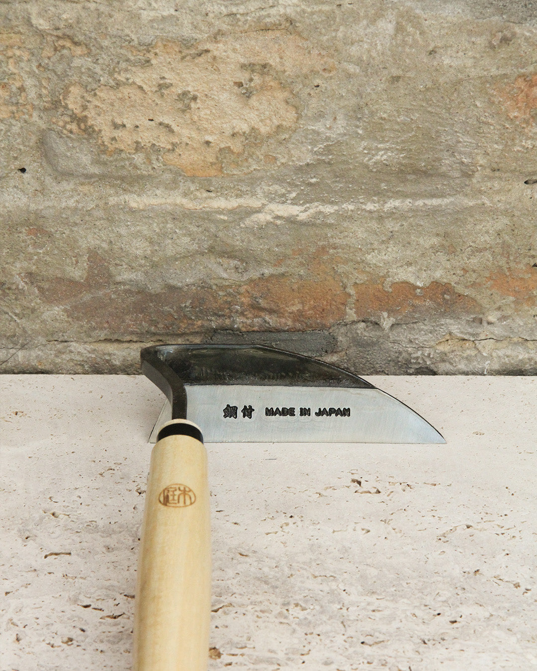 Die Niwaki | 'Unkrauthacke' Jäthacke für Rechts- und Linkshänder, mit einem Holzgriff und einer Metallklinge mit der Aufschrift MADE IN JAPAN, ruht auf einer hellen Steinfläche vor einer strukturierten Steinwand.
