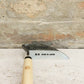 Eine Niwaki | „Weeding Hoe“ Jäthacke für Rechts- und Linkshänder von Niwaki mit Holzgriff und Metallklinge, auf der „MADE IN JAPAN“ sowie japanische Schriftzeichen eingraviert sind, ruht auf einer Steinoberfläche und lehnt an einer strukturierten Steinmauer – ideal zum Unkraut entfernen in Ihrem Garten.