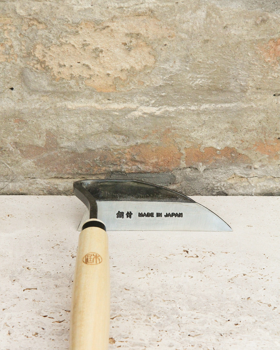 Eine Niwaki | „Weeding Hoe“ Jäthacke für Rechts- und Linkshänder von Niwaki mit Holzgriff und Metallklinge, auf der „MADE IN JAPAN“ sowie japanische Schriftzeichen eingraviert sind, ruht auf einer Steinoberfläche und lehnt an einer strukturierten Steinmauer – ideal zum Unkraut entfernen in Ihrem Garten.