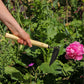 Die Niwaki | 'Unkrauthacke' Jäthacke für Rechts- und Linkshänder mit Holzgriff und schwarzer Klinge wird in der Nähe von grünen Pflanzen und blühenden rosa Blumen zum Entfernen von Unkraut im Garten eingesetzt.