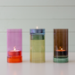 Drei farbenfrohe Glaskerzenhalter von Remember – das Windlicht „Lys“ in Rosa sowie Varianten in Gelb und Braun –, in denen jeweils eine brennende Kerze steht, sind nebeneinander auf einer weißen Fläche vor einem hellen Holzhintergrund angeordnet. Mit ihrem eleganten Color-Blocking-Design verleiht jedes mundgeblasene Windlicht von Remember jeder Umgebung einen lebendigen, handgefertigten Charme.