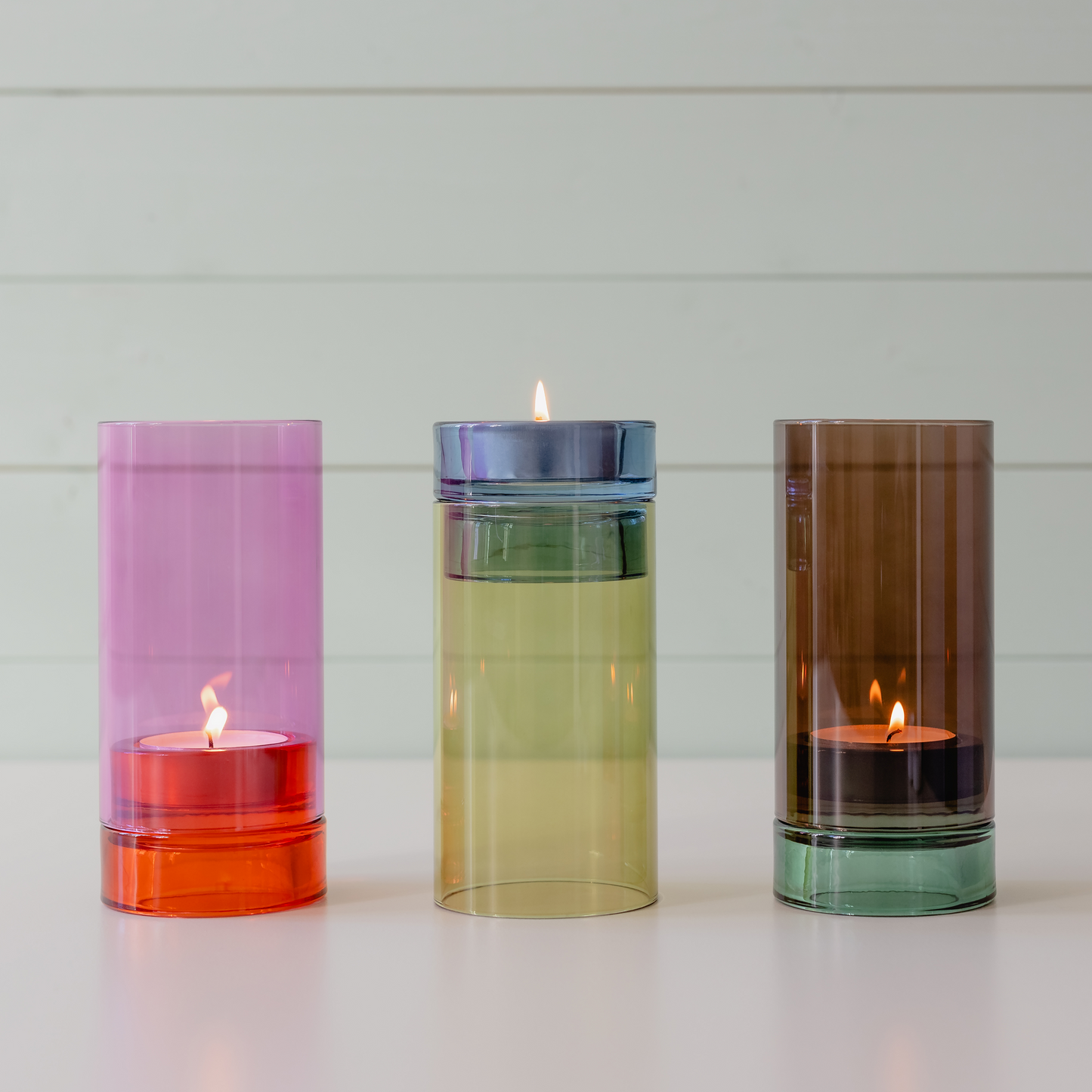 Drei farbenfrohe Glaskerzenhalter von Remember – das Windlicht „Lys“ in Rosa sowie Varianten in Gelb und Braun –, in denen jeweils eine brennende Kerze steht, sind nebeneinander auf einer weißen Fläche vor einem hellen Holzhintergrund angeordnet. Mit ihrem eleganten Color-Blocking-Design verleiht jedes mundgeblasene Windlicht von Remember jeder Umgebung einen lebendigen, handgefertigten Charme.