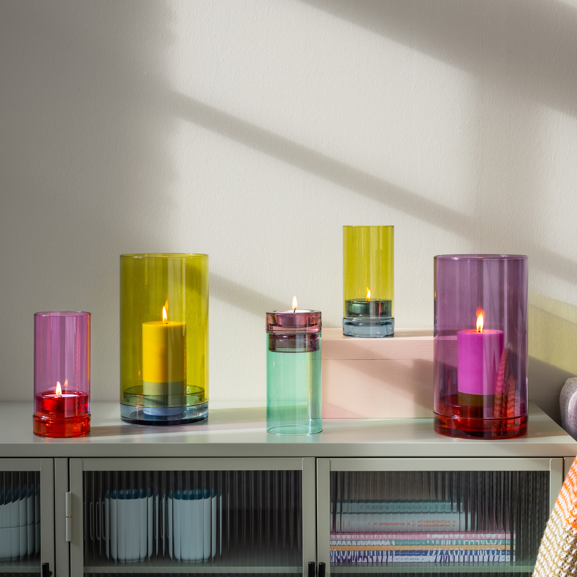 Ein „Lys“-Glaskerzenhalter der Marke Remember von Remember Windlicht mit leuchtend gelben, rosa und violetten Kerzen steht auf einem weißen Schrank in einem sonnendurchfluteten Raum. Licht und Schatten schaffen eine gemütliche, einladende Atmosphäre, während im Schrank darunter Tassen zu sehen sind.